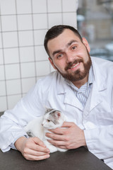 Vertical shot of a cheerful male veterinarian smiling to the camera, petting lovely white kitty on examination table