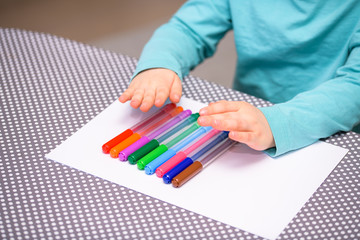 Close-up of five year old boy playing and lining up color pens on a white paper placed on a table with white dots. The boy is dressed in a turquoise shirt holding his hands above the pens.