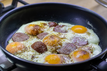 Fried eggs with sausage pepperoni and seasonings in a black pan on a gas stove. Tasty and nutritious yummy breakfast. Selective focus. Closeup view