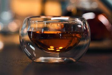 Transparent bowl with black tea on a wooden table. Blurred background of bright lights. Horizontal picture.