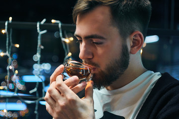 A young bearded guy is drinking black tea, holding a transparent bowl in his hands. Bright lights of a garland in the background.