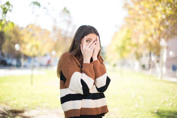 Anxiety - a conceptual image of a beautiful young caucasian woman covering her face with her hands and peering out with one eye between her fingers standing indoors. Scared from something or someone.