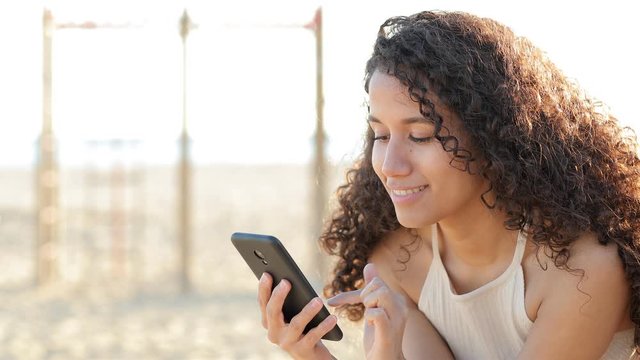 Happy latin woman using smart phone sitting on the beach