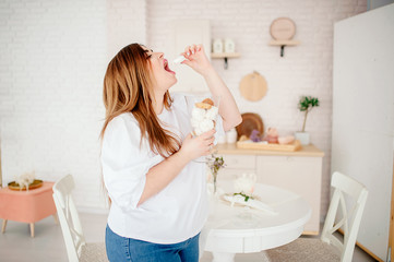 Happy plus size model in jeans and a white T-shirt, holding a sweet in her hands, opens her mouth in a stylish bright kitchen. The body is positive. The concept of one beauty. XXXL fashion