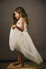 A young girl on gray backdrop in a white dress lifts her dress