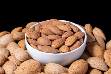 Peeled almonds in a white bowl surrounded by raw almonds in shell. Isolated on black background.