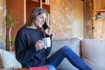 Serious young bruentte woman talking on the mobile sitting on the sofa at home drinking a tea wearing black sweater and blue jeans