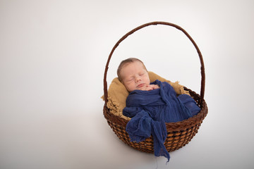Small baby new born swaddled in blue fabric laying asleep in brown wicker basket in studio portrait