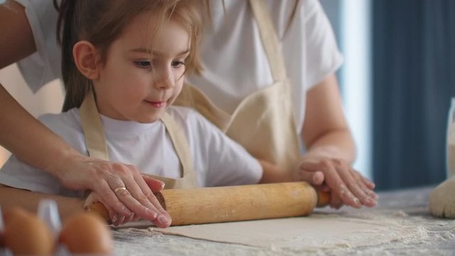 Mother Helping Daughter Roll Dough in Kitchen to Bake Cookies. Mom and daughter bake pizza in the kitchen together. Girl helps her mom to roll out the dough with a rolling pin. 