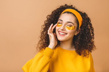 Image of young pure beautiful curly woman isolated over beige background take care of her skin with under eye patches.