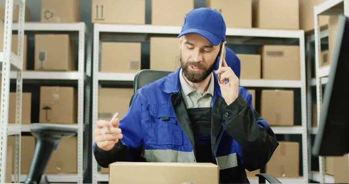 Caucasian postman talking on mobile phone while working at post office store with parcel at table. Man registering box and writing or marking its top while speaking on cellphone in mail storage.