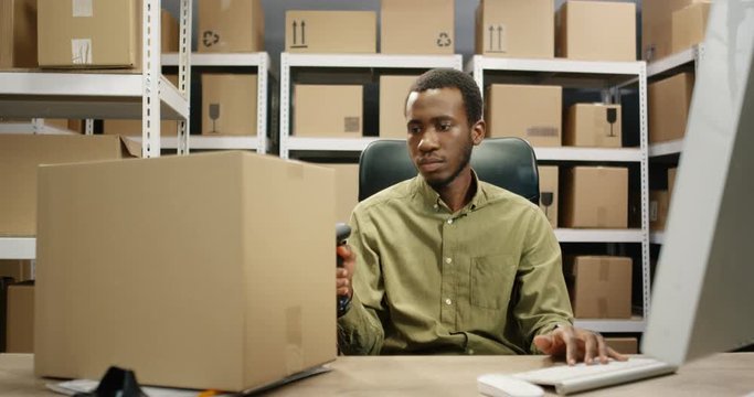 African American postman working at computer in post office store. Postman scanning bar code with scanner, registering parcel and filling in invoice document while sitting at desk.