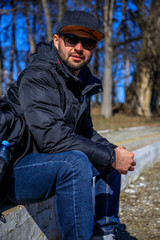 Stylish young man with a cap, sunglasses, sneakers in a denim jacket with a backpack sits on golden foliage in the park. Attractive hipster guy is rests at the forest.