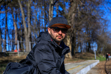 Stylish young man with a cap, sunglasses, sneakers in a denim jacket with a backpack sits on golden foliage in the park. Attractive hipster guy is rests at the forest.