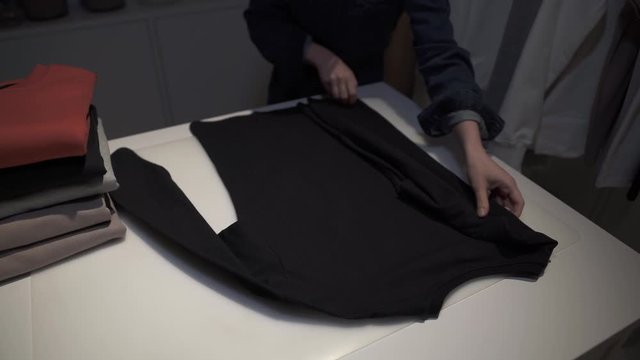 Handheld shot of woman folding a black sweatshirt on a table in a showroom. Close up of female hand folding a grey pullover sweatshirt on a white table
