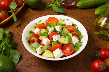 Salad with tomato, avocado, cucumber and  feta cheese in a white bowl on a rustic wooden table. Selected focus.