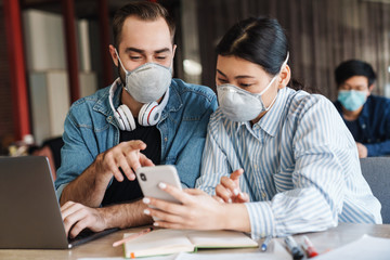 Photo of young students in medical masks using cellphone while studying