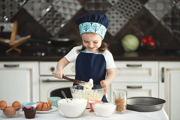 A cute little girl in an apron and a Chef's hat is stirring the dough with a wooden spatula, looking at the camera and smiling while cooking.