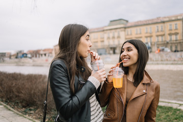 Two pretty girls drinking juice with straws next to river