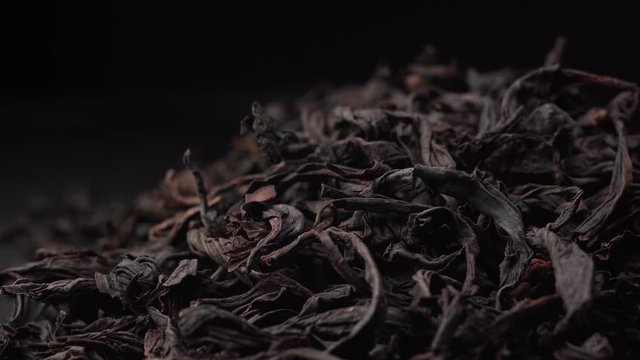 leaves of fragrant black tea revolve on black table. Beautiful green tea lies on table and camera rotates around it.