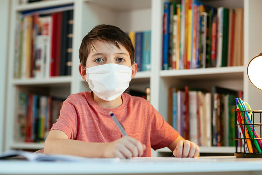 Young student wearing protective mask working in isolation on school assignments. Front view of child wearing face mask studying at home during coronavirus outbreak.