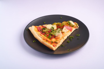 Pizza slice with pepperoni, salami, melted mozzarella cheese, pickles and dill in black plate on white background, angle view