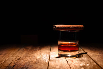 Whiskey and cigar on the wooden weathered table. Handmade cuban cigar lies on a glass.