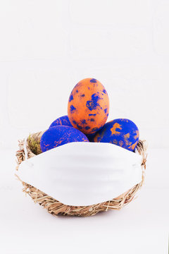 Easter Time In Quarantine Concept. Face Mask On A Nest With With Classic Blue And Orange Speckled Easter Eggs On The Table On A White Breakwall Background