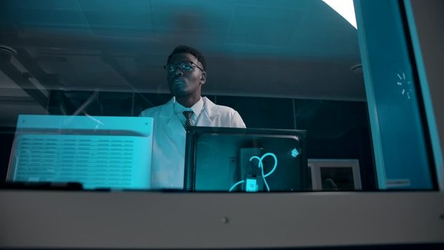 The middle view of a black skin doctor in a white robe overseeing surgery from the control room. Few monitors with surgery data for helping his colleagues.