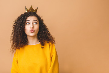 Gorgeous beautiful girl with curly brown hair and wearing casual and holding crown on head isolated over beige studio background. Sending air kiss.