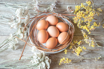 Brown raw eggs on a plate with white and yellow flowers on wooden table. Easter composition, mockup on rustic background. 