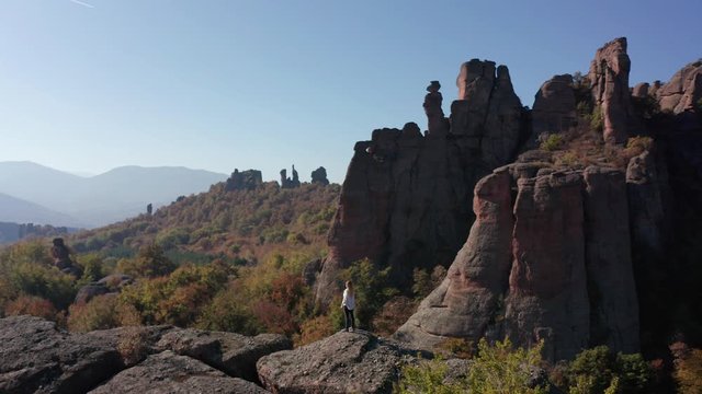 Aerial fly around a young blonde woman with white shirt feeling freedom while standing at the edge of stunning rock formation surrounded by autumn forest, Belogradchik Rocks, Bulgaria.