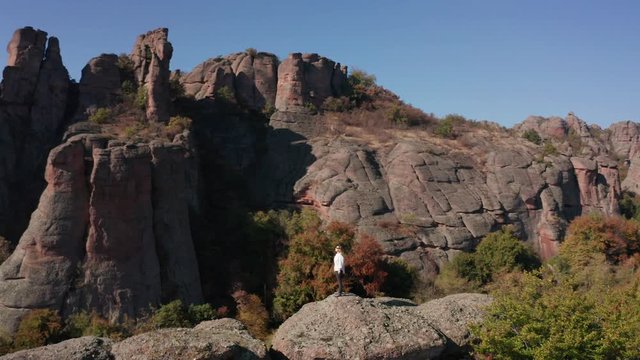 Aerial fly around a young blonde woman with white shirt feeling freedom while standing at the edge of stunning rock formation surrounded by autumn forest, Belogradchik Rocks, Bulgaria.