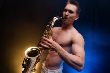 Muscular man with naked torso playing on saxophone with smoked colorful background 