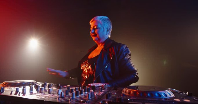 Funny grandma is a dj. Authentic mature woman in cool outfit working at turntables in a nightclub, rocking the party up 4k footage