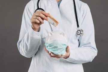 Doctor with Stethoscope Holding Piggy Bank Wearing Medical Face Mask. Coronavirus concept