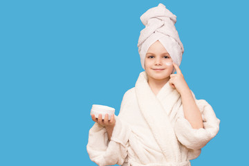 Caucasian child girl model with towel on her head puts cream on her face touching under eyes isolated on blue background.