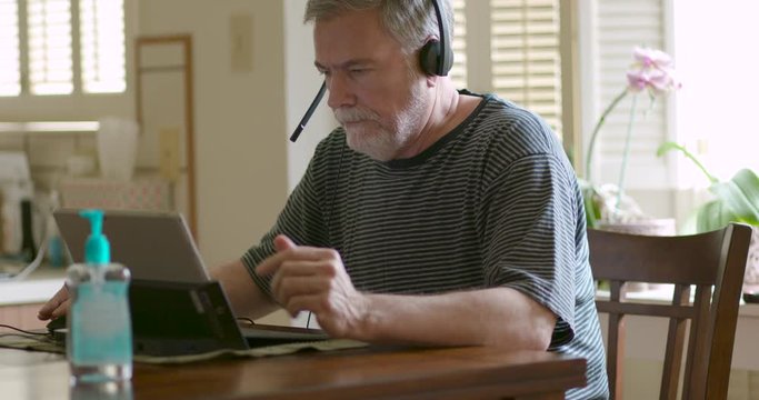 A mature Caucasian male wearing a headset with a microphone and using a laptop as he telecommutes to a meeting from home because of mandates created by the spread of the coronavirus COVID19.