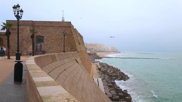 The rainy morning on Atlantic Ocean shore with a view on Baluarte de San Roque (bastion) of Cadiz fortress, foggy rainy sky and stormy waters, Spain