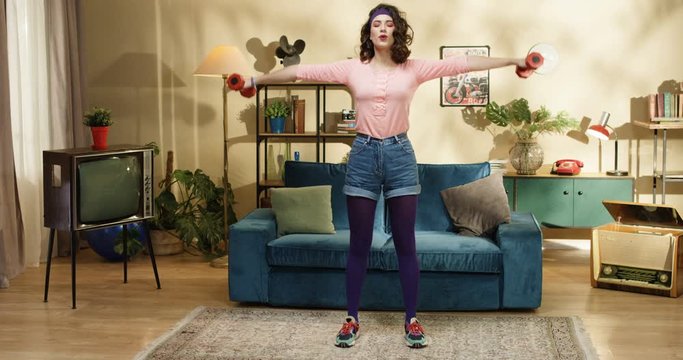 Funny young pretty stylish female doing exercise on biceps with red dumbbells in living room. Retro style fitness Caucasian girl having home workout with equipment indoor 60s healthy lifestyle concept