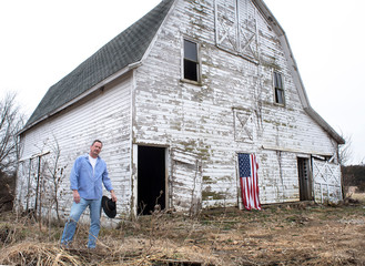 Man with cowboy hat standing in front of old abandoned barn in country, American flag hanging on barn © Jon