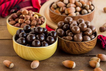 Milky and dark chocolate covered hazelnuts in bamboo bowls on wooden table with natural hazelnuts.
