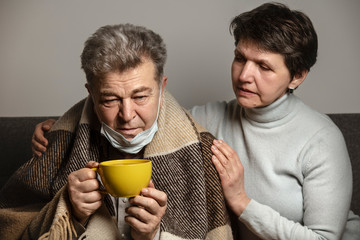 A couple sitting on a sofa on a quarantine at home. An sick older man with protective mask drink a tea to protect against virus. Family support for the illness. Coronavirus. COVID-19.