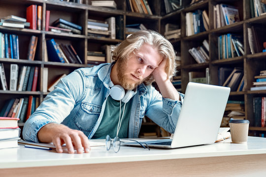 Unhappy bored lazy tired male college university student studying in library looking at laptop feel fatigue disinterested in dull tedious learning online class preparing for exam in library concept.