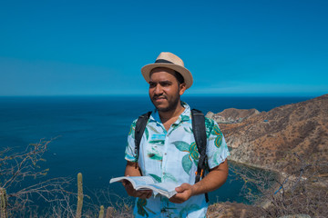man checking travel guide on top of a mountain. sea in the background