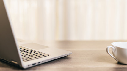 Laptop and white ceramic coffee cup on wooden desk in the morning, with white translucent shade in...