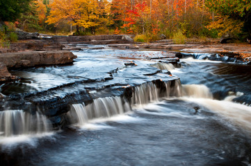 Fototapeta na wymiar The Sturgeon River spills over a ledge waterfall as it flows through a landscape of peak autumn color in Michigan's Upper Peninsula.
