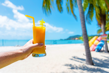First-person view. Girl holds a glass cup of cold mango fresh on the background of a sandy tropical beach. White sand and palm trees. Fairytale vacation