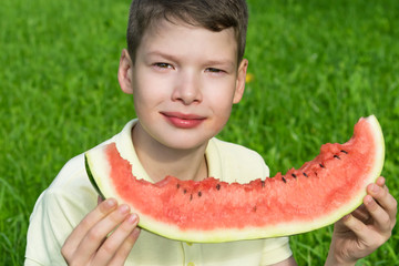 handsome boy eating a large slice of red watermelon, on a background of green grass