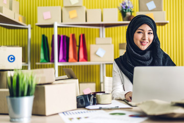 Portrait of smiling beautiful muslim owner asian woman freelancer sme business online shopping working on laptop computer with parcel box on table at home - Business online shipping and delivery